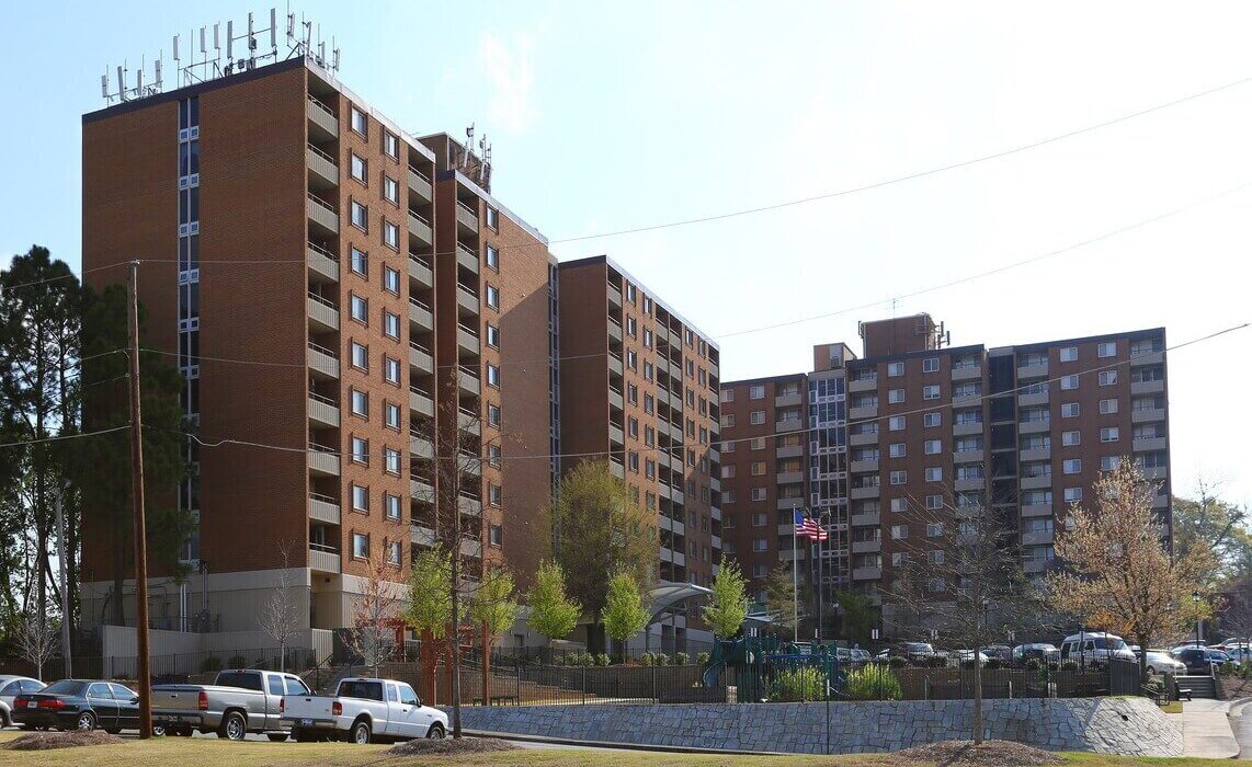 Exterior view of Cosby Spear Highrise, a multi-story senior residential building in Atlanta, Georgia, featuring a red brick facade with rows of windows and surrounded by trees under a clear blue sky.