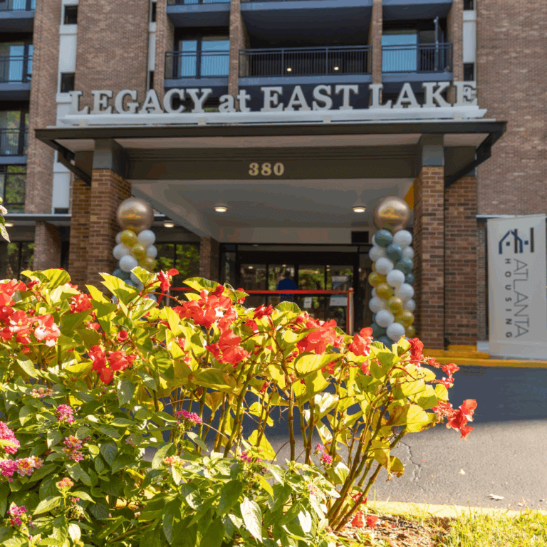 Front entrance of Legacy at East Lake, a brick residential building with a sign above the doorway, festive gold and white balloon columns, and colorful flowers in the foreground.